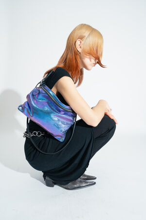 A person in black clothing poses in front of a white background with the two-tone metallic leather crossbody bag MOONLIT ed.1 in purple-blue 