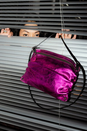 A person peeks through window blinds, where an INA KENT vibrant pink metallic bag is artfully draped.