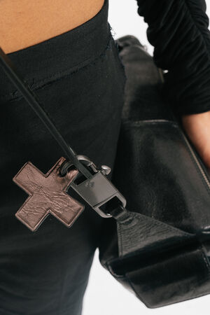 Nahaufnahme einer Person in schwarzen Hosen und einem schwarzen Oberteil, die eine schwarze Ledertasche von INA KENT mit einem kreuzförmigen Lederschlüsselanhänger am Riemen trägt.
