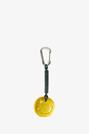 A keychain by INA KENT with a metal carabiner, black strap, and a gold smiley face charm hanging at the end.