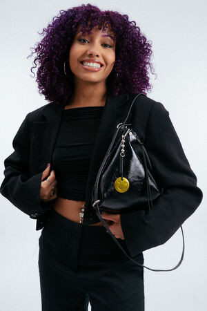 Frau mit lockigem lila Haar lächelt, während sie einen schwarzen Blazer und ein bauchfreies Top trägt und eine glänzende schwarze Handtasche von INA KENT hält, die mit einem Schlüsselanhänger mit einem gelben Smiley verziert ist.