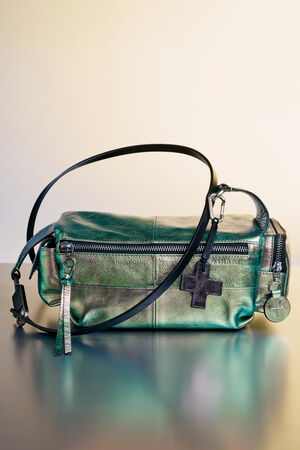 A metallic green INA KENT leather crossbody bag with a black strap, zipper closure, and black cross and round keychain charms, placed on a reflective surface.