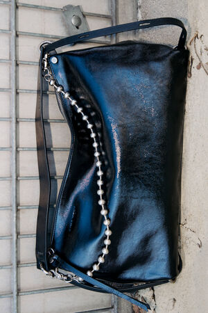 A black leather INA KENT handbag with a shiny finish, black strap, and decorative string of silver beads sits on a concrete surface beside a metal grate.