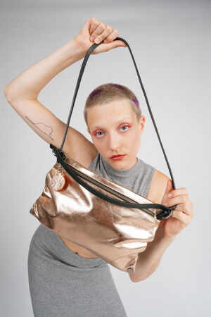Person with short hair holding an INA KENT metallic bag by the strap, posing against a plain background, wearing a sleeveless gray top.