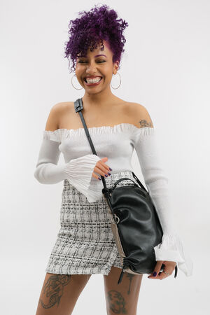 Woman with curly purple hair, wearing a white off-shoulder top and plaid skirt, smiling while holding a chic black INA KENT bag, standing against a plain white background.