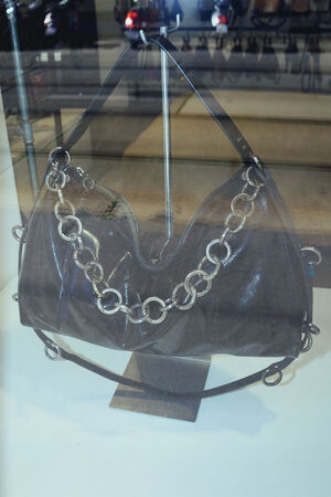 Eine dunkle Lederhandtasche von INA KENT mit einem klobigen silbernen Kettenriemen wird auf einem Ständer hinter einem Glasfenster präsentiert.