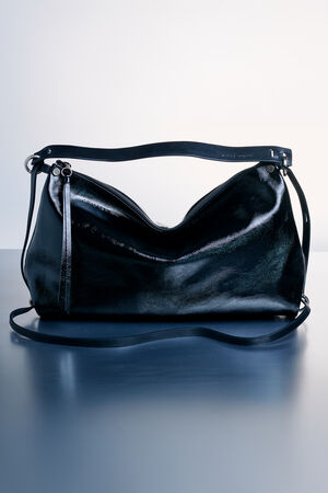 An INA KENT black leather shoulder bag with a shiny finish, silver hardware, and two adjustable straps sits on a blue surface against a light background.