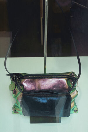 Eine zweifarbige Handtasche von INA KENT mit einem schillernden grün-rosa Korpus und einer schwarzen Vordertasche wird mit einem dünnen Schulterriemen auf einer weißen Oberfläche präsentiert.