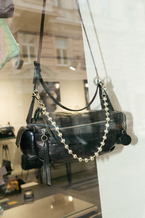 Eine schwarze Lederhandtasche von INA KENT mit Reißverschluss, dekorativem Kettenriemen und Schlüsselanhänger hängt in einem Schaufenster.