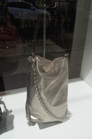 Eine metallisch-silberne INA KENT-Umhängetasche mit perlenbesetztem Riemen wird auf einer weißen Fläche in einem Schaufenster präsentiert.