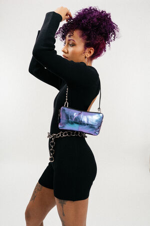 A person with curly purple hair wears a black long-sleeve outfit and carries an INA KENT metallic purple purse with a chain strap around their waist.