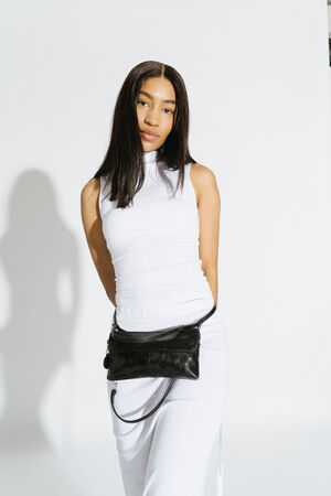 Woman in a sleeveless white dress stands against a plain white background, with an INA KENT black bag worn crossbody at her waist.