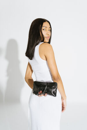 Woman in a white sleeveless dress stands facing away, looking over her shoulder, holding a small black INA KENT leather bag behind her back.
