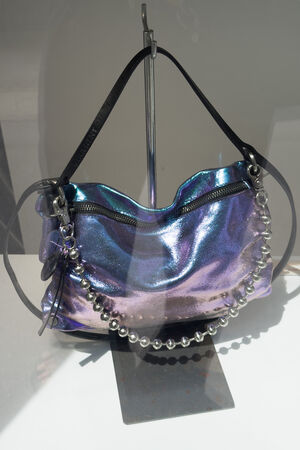 Eine glänzende, schillernde, violette Handtasche von INA KENT mit schwarzen Riemen, Reißverschluss und einer dekorativen silbernen Perlenkette, präsentiert auf einem Ständer.