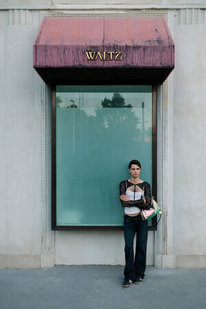A person stands in front of a building with a large window and a faded red awning labeled "WALTZ," holding an INA KENT bag and wearing dark pants and a black lace top.