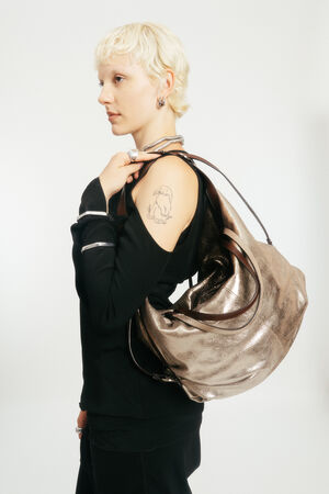 Person with short blond hair wearing a black outfit and carrying a large metallic INA KENT shoulder bag; a tattoo of a bear on their upper arm is visible.