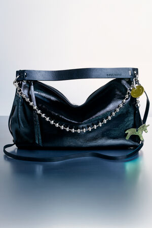 A black leather INA KENT handbag with a silver bead chain, a green animal-shaped charm, and a round gold tag, displayed on a blue surface.