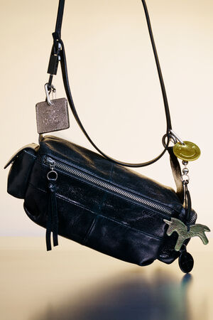 A black leather INA KENT crossbody bag with a front zipper, decorative tags, and a green horse-shaped keychain, displayed against a neutral background.