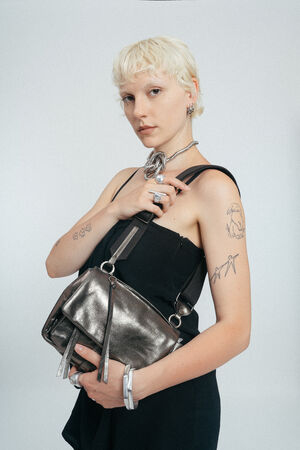 Person with short blonde hair in a black dress poses with an INA KENT metallic silver handbag, showing several tattoos and wearing chunky silver jewelry against a plain background.