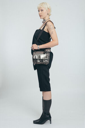 Person with short blonde hair stands in profile wearing a black sleeveless outfit, black boots, a metallic INA KENT shoulder bag, and silver accessories, with visible tattoos on their left arm.
