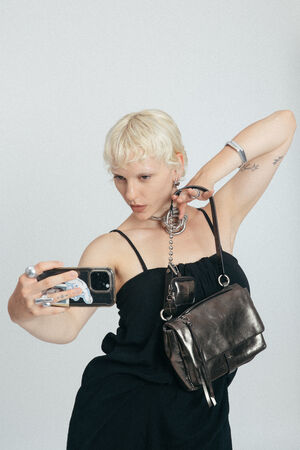 Person with short blonde hair takes a selfie, wearing a black dress and holding an INA KENT metallic purse over their shoulder. Their arm is bent behind their head, displaying tattoos and jewelry.