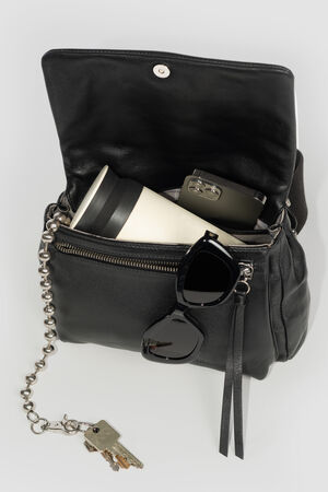 An INA KENT black leather handbag with sunglasses, a reusable coffee cup, and a smartphone inside. Keys on a chain are attached to the bag.