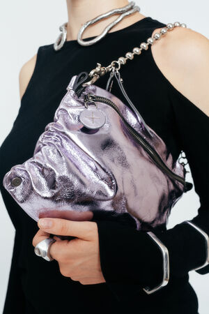 A person wearing a black, cut-out shoulder top holds an INA KENT metallic purple, textured handbag with a chain strap and a round charm.