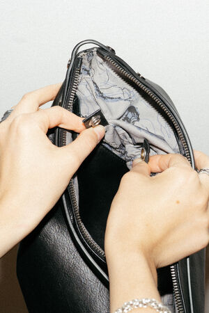 Hands opening an INA KENT black leather bag to reveal a light-colored, patterned interior lining and metal hooks.