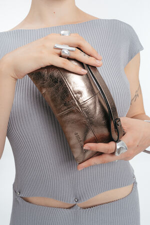 A person wearing a grey ribbed dress and silver rings holds an INA KENT metallic bronze clutch bag with a zipper.