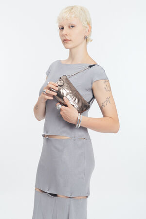 A person with short blond hair in a light gray dress holds an INA KENT metallic handbag. They have visible tattoos, several rings and bracelets, all set against a plain white background.
