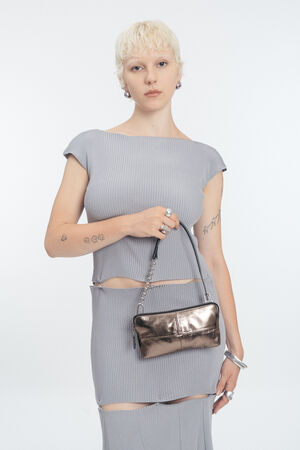 Person with short blond hair wearing a ribbed gray outfit, holding an INA KENT metallic purse, standing against a plain white background.