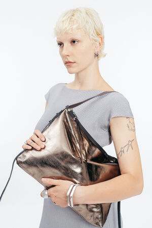 Person with short blond hair wearing a gray top holds a large metallic bronze INA KENT bag; visible bird tattoos on left arm and silver jewelry on fingers and wrist.