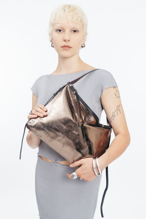 Person with short blonde hair wearing a grey ribbed dress holds a large metallic INA KENT handbag, displaying several tattoos, rings, and bracelets, against a plain white background.
