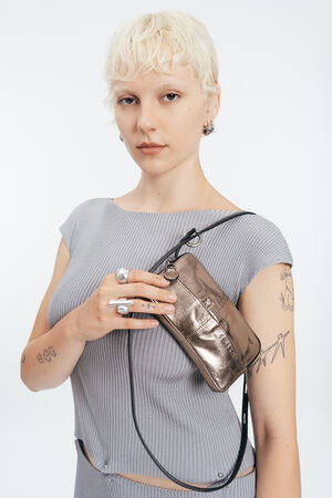 Person with short blonde hair and visible tattoos wears a light grey ribbed top, holding an INA KENT metallic purse with a shoulder strap across their chest.