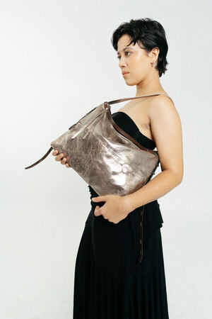 A person in a black dress stands holding a large metallic INA KENT handbag under their arm against a plain white background.
