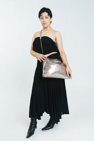 Person wearing a strapless black dress and black boots stands while holding an INA KENT metallic crossbody bag against a plain light background.