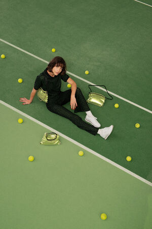 Eine Person in dunklem Outfit und weißen Turnschuhen sitzt auf einem grünen Tennisplatz, umgeben von gelben Tennisbällen und goldfarbenen INA KENT-Taschen.