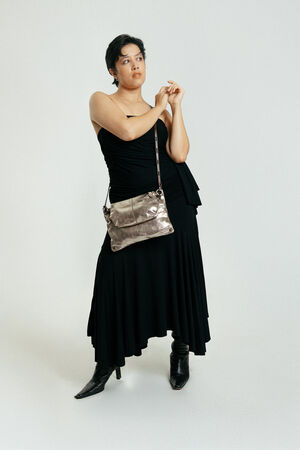 Person wearing a one-shoulder black dress, black boots, and holding an INA KENT metallic crossbody bag stands against a plain white background.