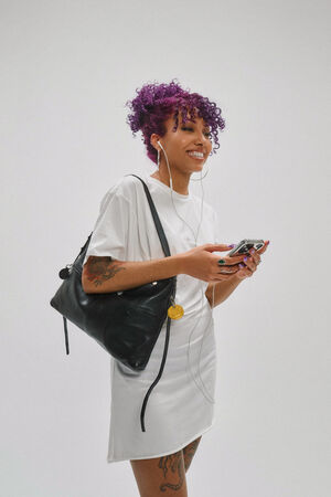 A person with curly purple hair, wearing a white dress and an INA KENT black bag, holds a phone and smiles while listening to earphones. Visible tattoos adorn their arms and legs.