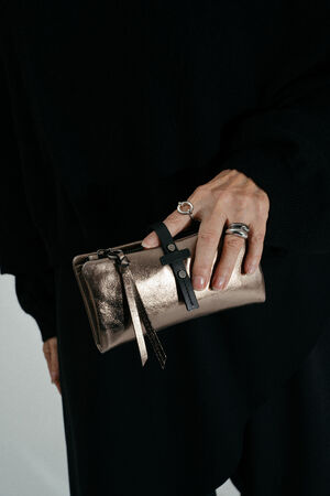 A person wearing silver rings holds a small metallic INA KENT clutch purse with black strap detail, dressed in a sleek black outfit.