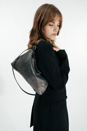 A woman in a black blazer stands sideways, looking down, with an INA KENT metallic shoulder bag slung over her left shoulder against a plain white background.