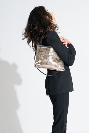 A woman in a black suit stands in profile, holding an INA KENT metallic shoulder bag with both hands against a plain white background.