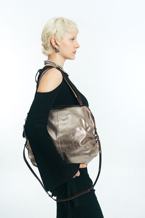 A person with short blonde hair wears a black cold-shoulder top and black pants, carrying a large metallic INA KENT tote bag with brown straps over their shoulder.