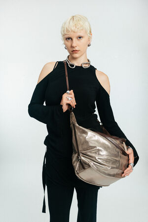 Person with short blond hair in a black cold-shoulder top and black pants holds a large metallic gold INA KENT shoulder bag against a plain light background.