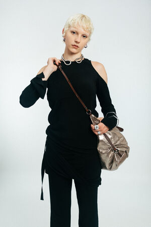 Person with short blonde hair wearing a black outfit and silver accessories, holding an INA KENT metallic shoulder bag, standing against a plain light background.