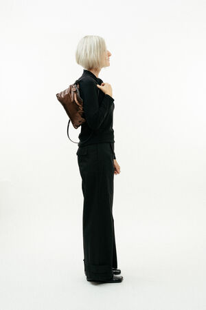 Older woman with short gray hair, dressed in black, stands in profile against a white background, wearing an INA KENT brown backpack over one shoulder.