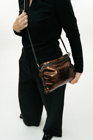 Person dressed in black holds an INA KENT metallic brown crossbody bag with a zipper and round charm, standing against a plain white background.