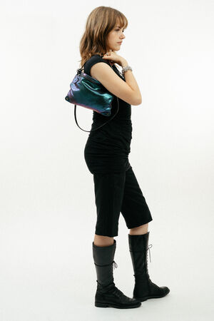 A person stands in profile wearing a black top, knee-length black shorts, black knee-high boots, and carrying a small iridescent INA KENT shoulder bag.
