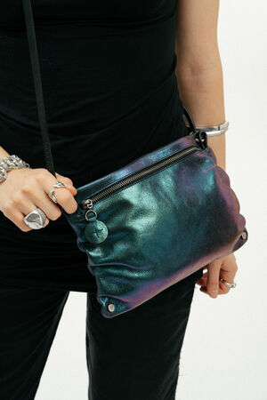 Person wearing a black outfit holds an INA KENT metallic, iridescent crossbody bag with a zipper and round keychain, accessorized with silver rings and bracelets.