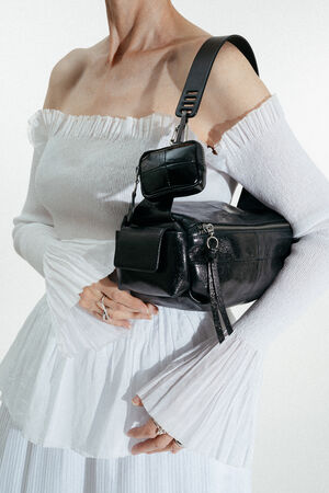 A person in an off-the-shoulder white dress holds a large black INA KENT leather bag with a smaller pouch attached to the strap.